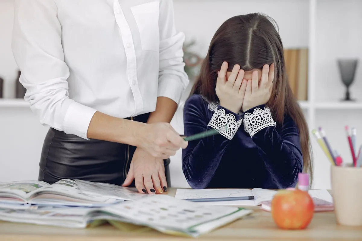 Young student in blue velvet dress covering face with hands in frustration while adult in white shirt provides support during study session with open textbooks on desk