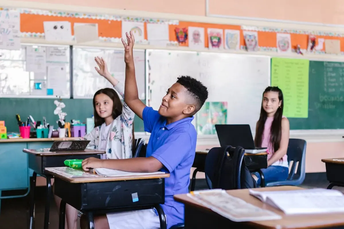 3 children in a classroom; a boy in blue tshirt raising his hand